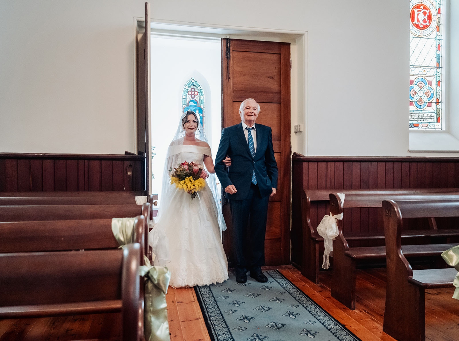 keyneton soldier's memorial hall wedding - lara & simon 4 Bride Lara in an off-the-shoulder wedding gown enters Keyneton Church arm in arm with an older man, carrying a colourful bouquet.