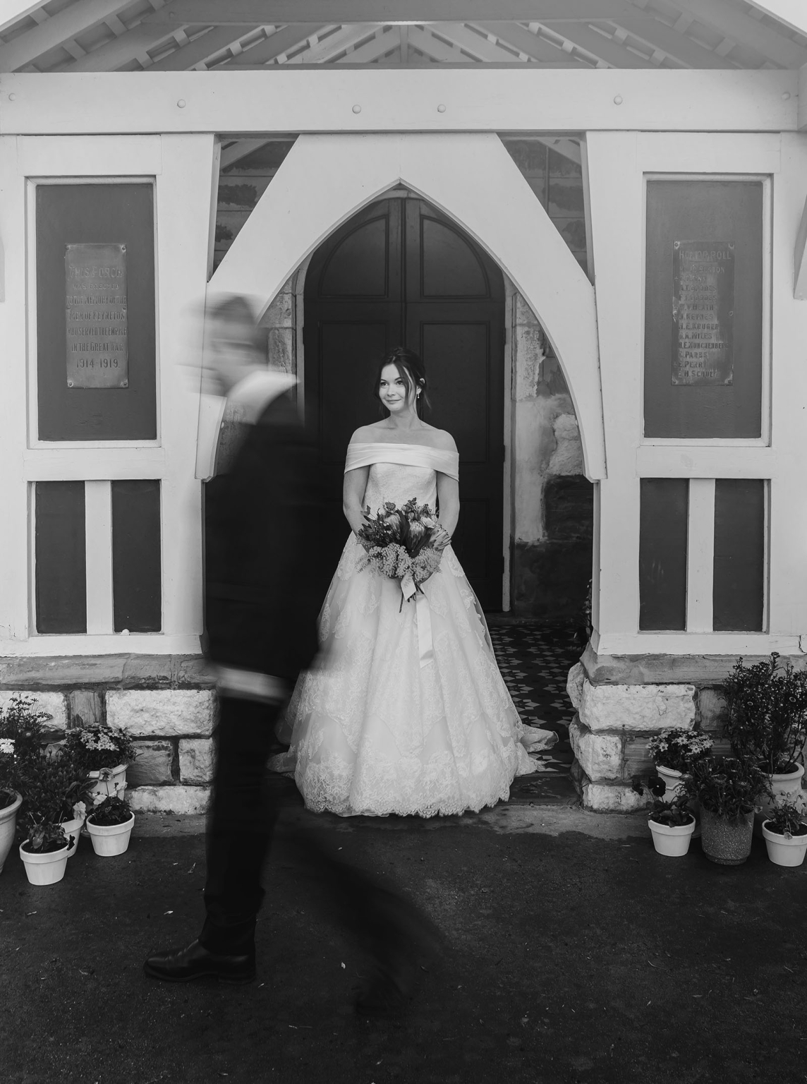 keyneton soldier's memorial hall wedding - lara & simon 32 Bride Lara holding a bouquet in the arched doorway of Keyneton church as the groom passes in soft motion blur.