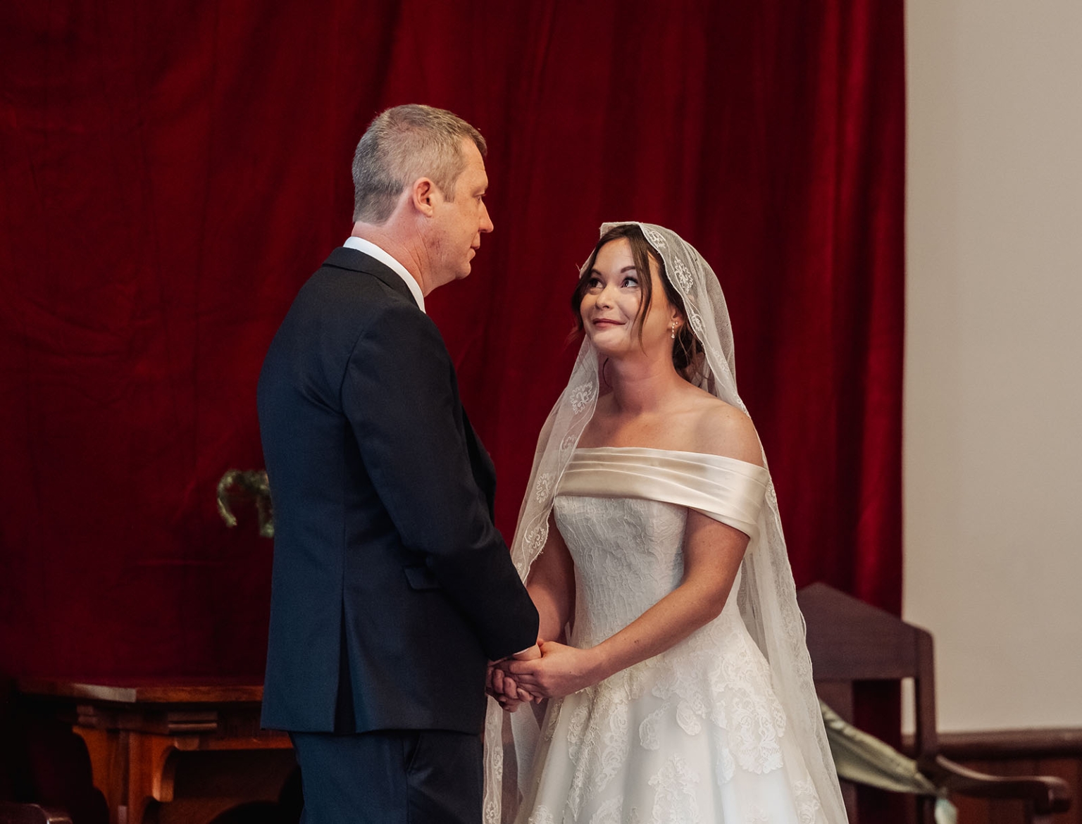 keyneton soldier's memorial hall wedding - lara & simon 10 Lara and Simon holding hands during their wedding ceremony, with red curtains behind them