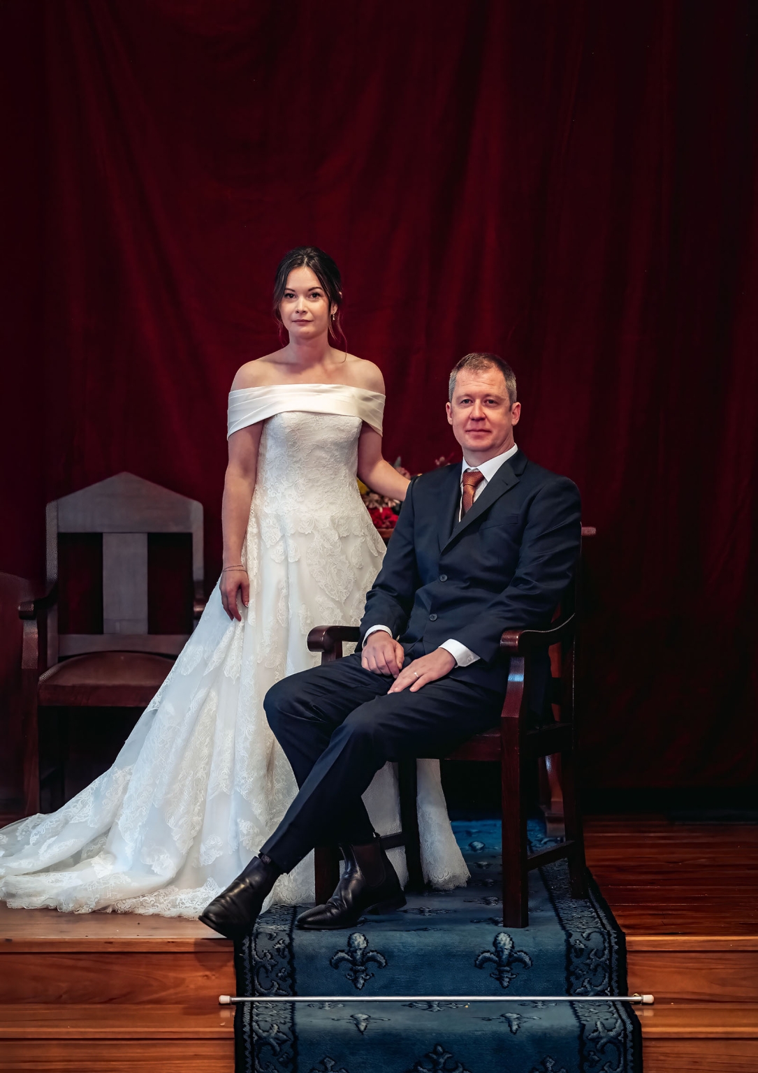 keyneton soldier's memorial hall wedding - lara & simon 27 Bride in a lace wedding gown standing beside groom seated in a wooden chair before a deep red curtain