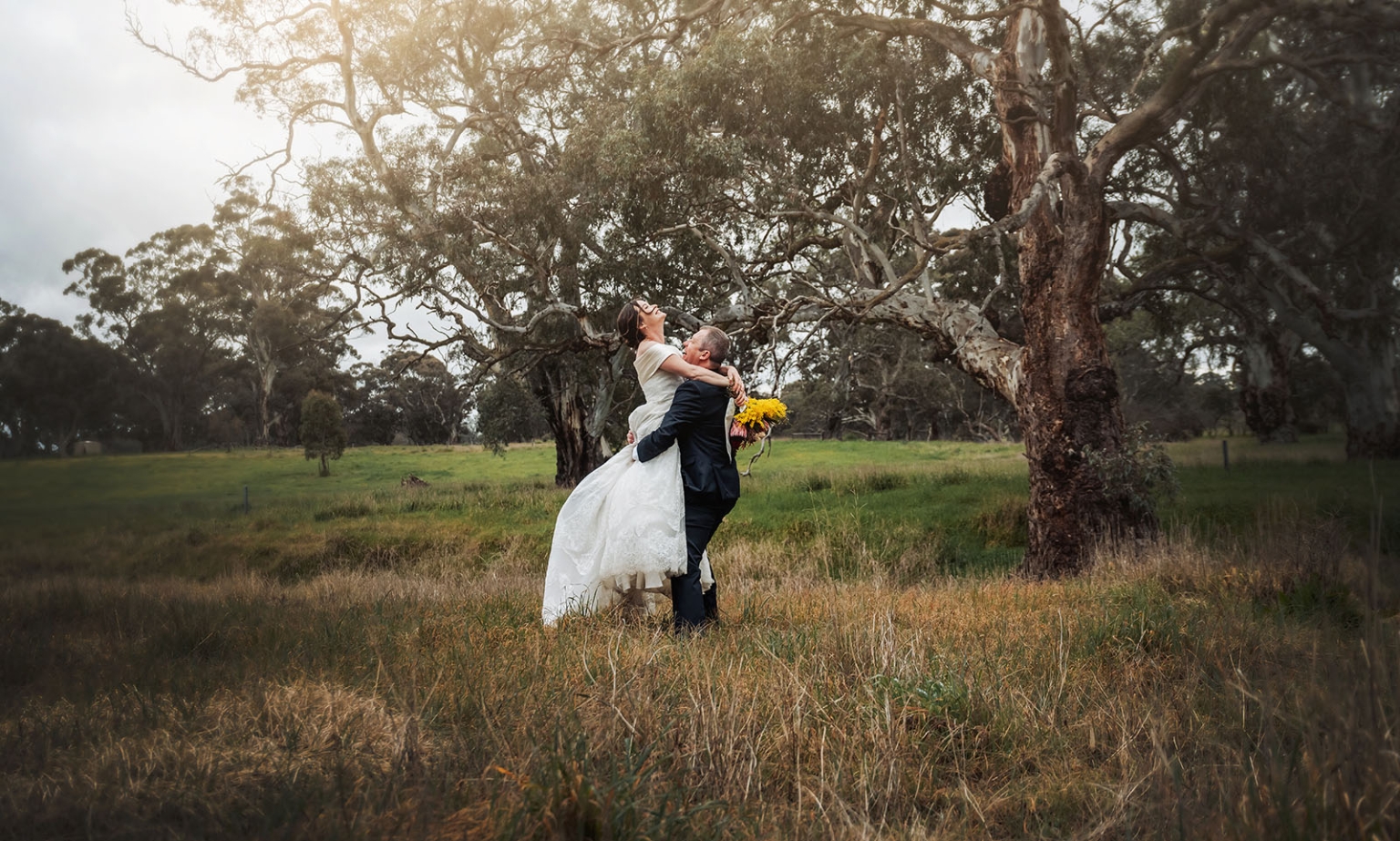 keyneton soldier's memorial hall wedding - lara & simon 40 Simon lifts Lara in a grassy field beneath large eucalyptus trees, with her yellow bouquet visible.