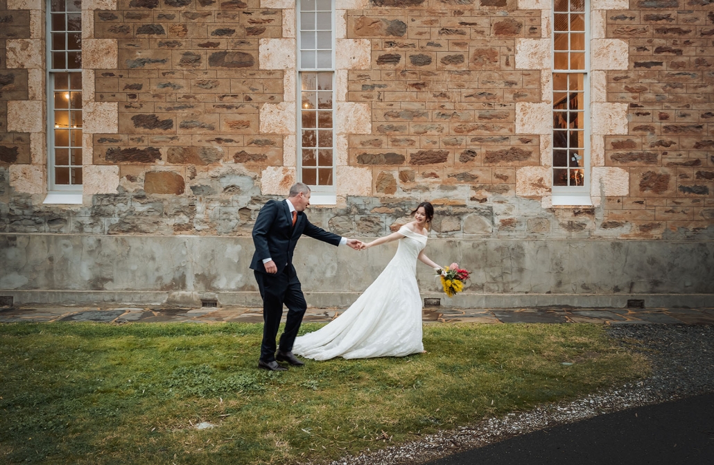 Lara and Simon holding hands outside the stone Keyneton church, with Lara carrying a colourful bouquet.