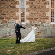 Lara and Simon holding hands outside the stone Keyneton church, with Lara carrying a colourful bouquet.