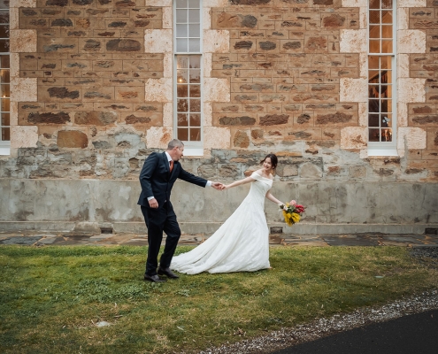 Lara and Simon holding hands outside the stone Keyneton church, with Lara carrying a colourful bouquet.