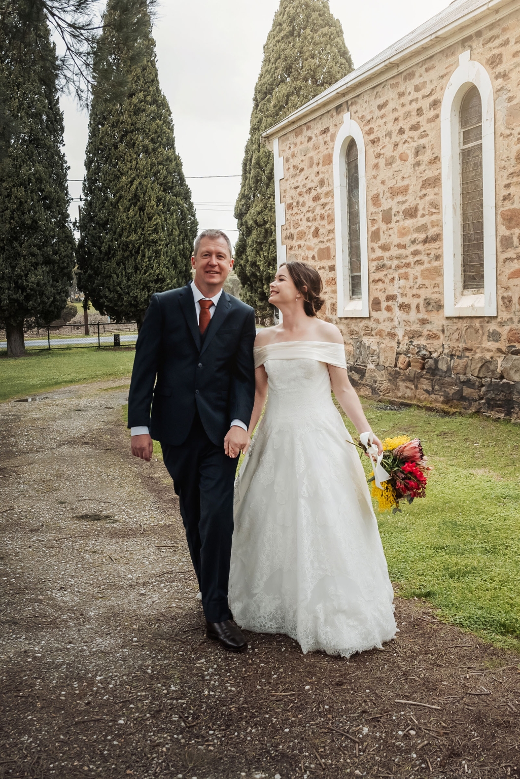 keyneton soldier's memorial hall wedding - lara & simon 24 Lara and Simon walking hand in hand beside the stone church at Keyneton, with the bride holding a colourful bouquet