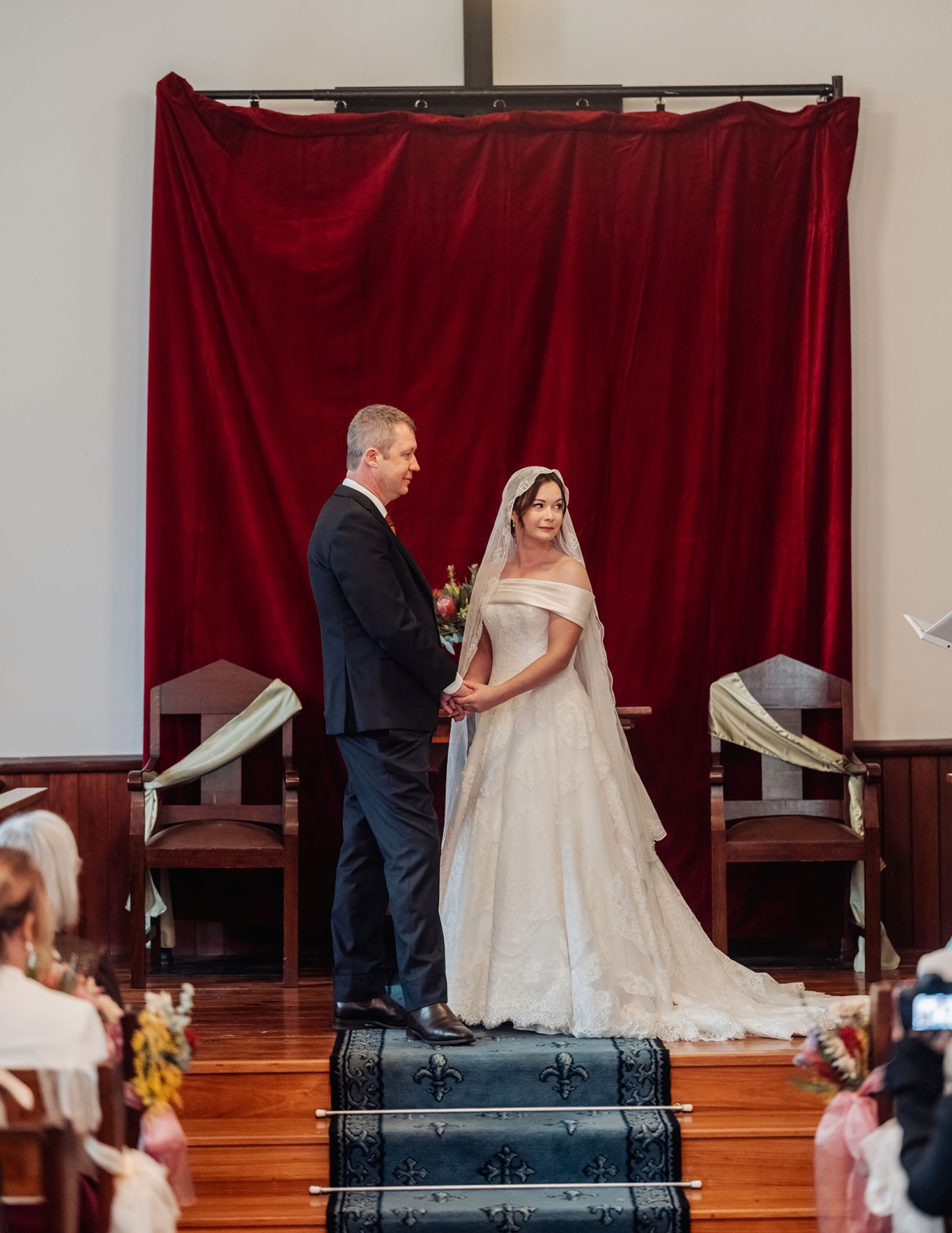 keyneton soldier's memorial hall wedding - lara & simon 6 Lara and Simon holding hands during their wedding ceremony before a deep red curtain in Keyneton church