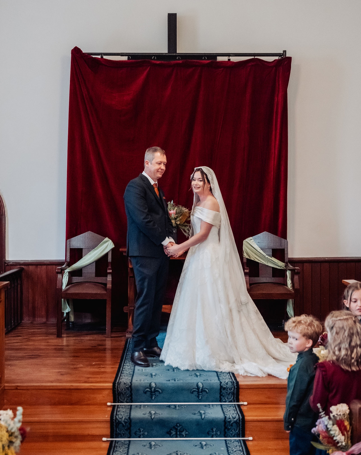 keyneton soldier's memorial hall wedding - lara & simon 9 Lara and Simon holding hands during their wedding ceremony in the Keyneton church, beneath a red velvet-covered cross
