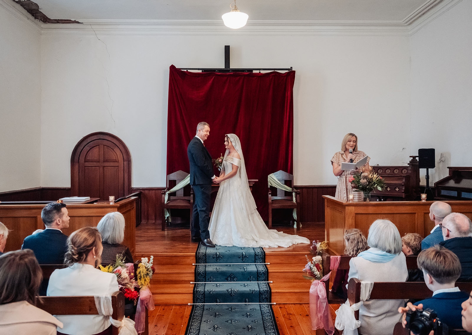 keyneton soldier's memorial hall wedding - lara & simon 12 Lara and Simon holding hands during their wedding ceremony in the Keyneton church, with guests seated in pews and their celebrant reading beside them.