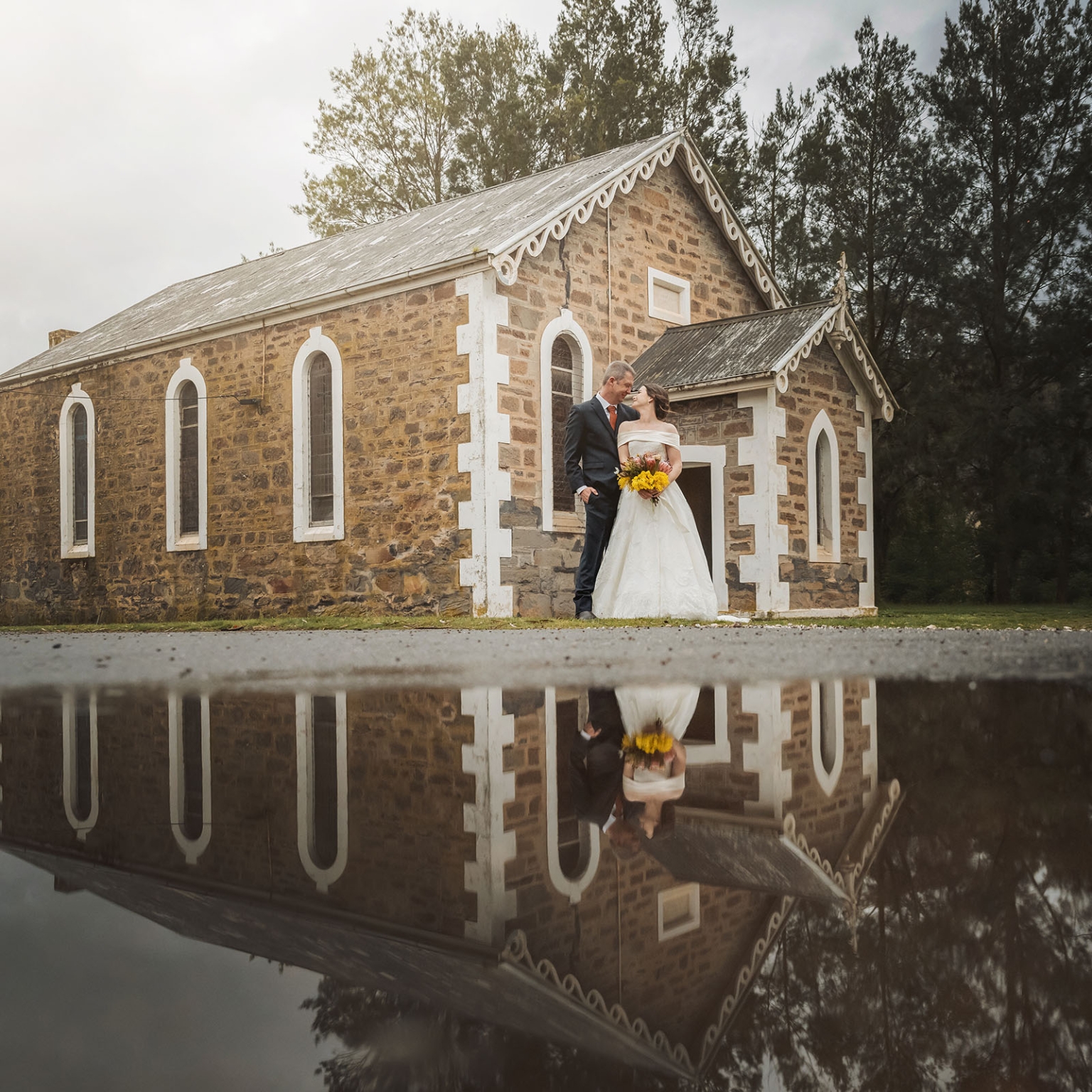keyneton soldier's memorial hall wedding - lara & simon 20 Lara and Simon standing together outside the stone Keyneton church, reflected in a puddle on the road