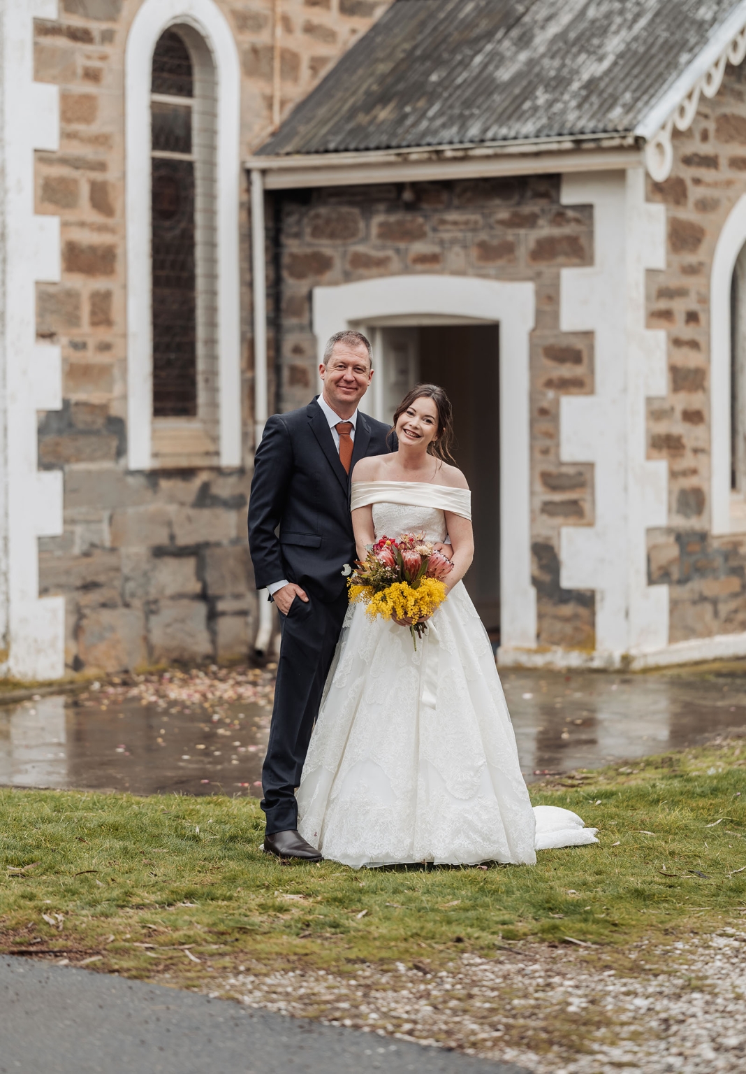 keyneton soldier's memorial hall wedding - lara & simon 19 Lara and Simon standing outside the stone church beside Keyneton Soldier's Memorial Hall, with Lara holding a bright native flower bouquet