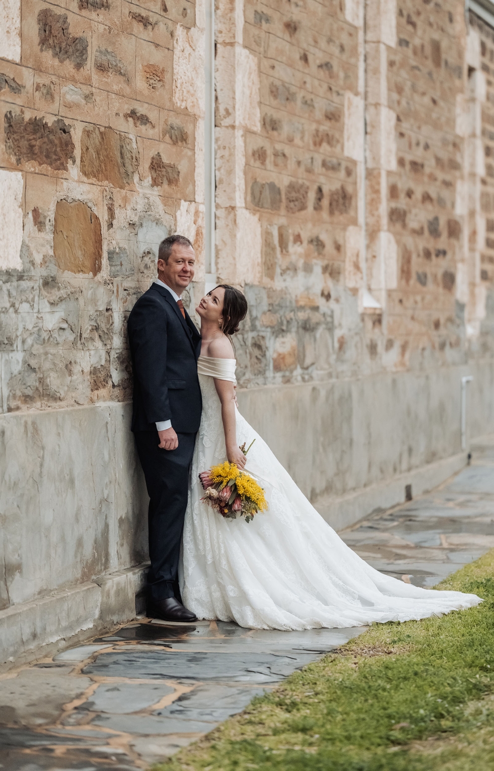 keyneton soldier's memorial hall wedding - lara & simon 34 Lara and Simon leaning against a textured stone wall, with Lara holding a yellow bouquet in her off-the-shoulder wedding gown.
