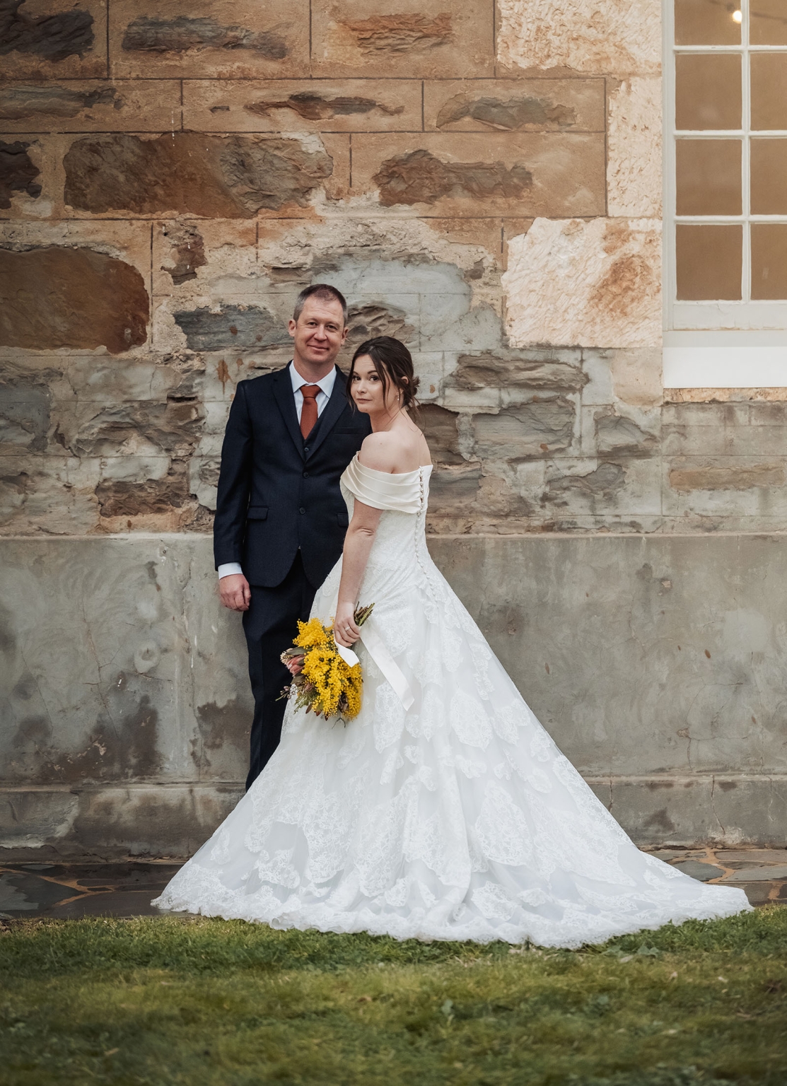 keyneton soldier's memorial hall wedding - lara & simon 33 Lara and Simon posing outside against a weathered stone wall, with Lara holding a yellow bouquet in her lace wedding gown.