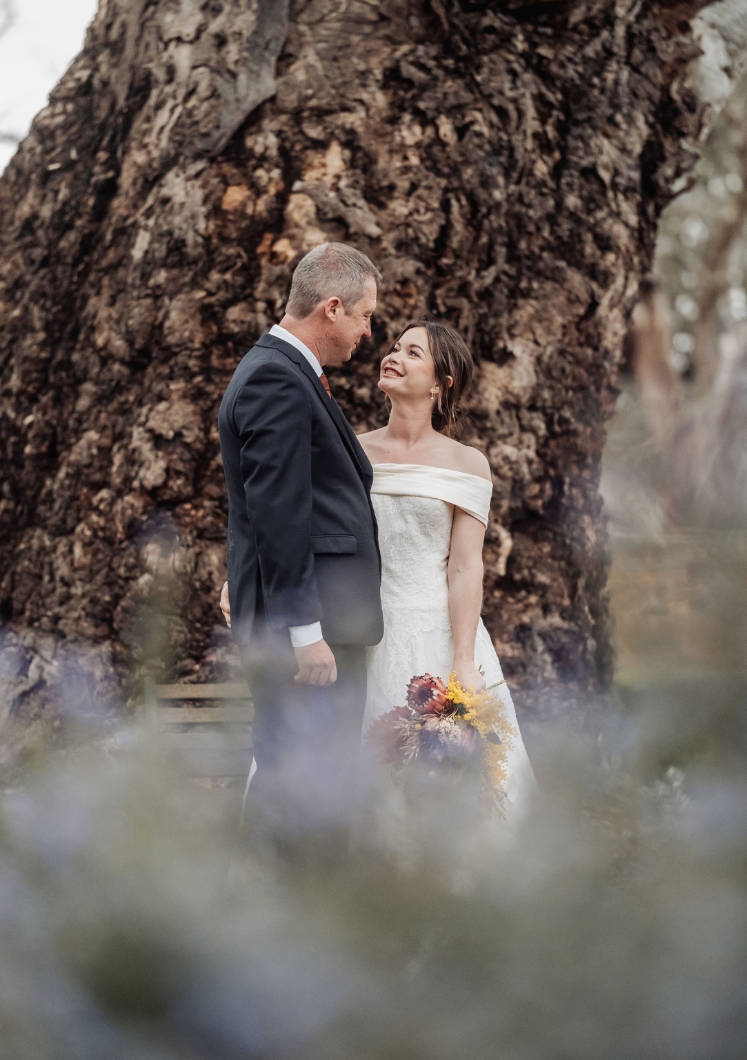 keyneton soldier's memorial hall wedding - lara & simon 37 Lara and Simon smiling at each other beside a large tree, with Lara holding a native flower bouquet.