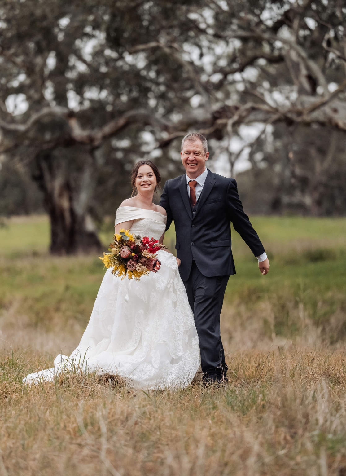 keyneton soldier's memorial hall wedding - lara & simon 38 Lara and Simon walking through dry grass beneath a large tree, with Lara holding a colourful bouquet.