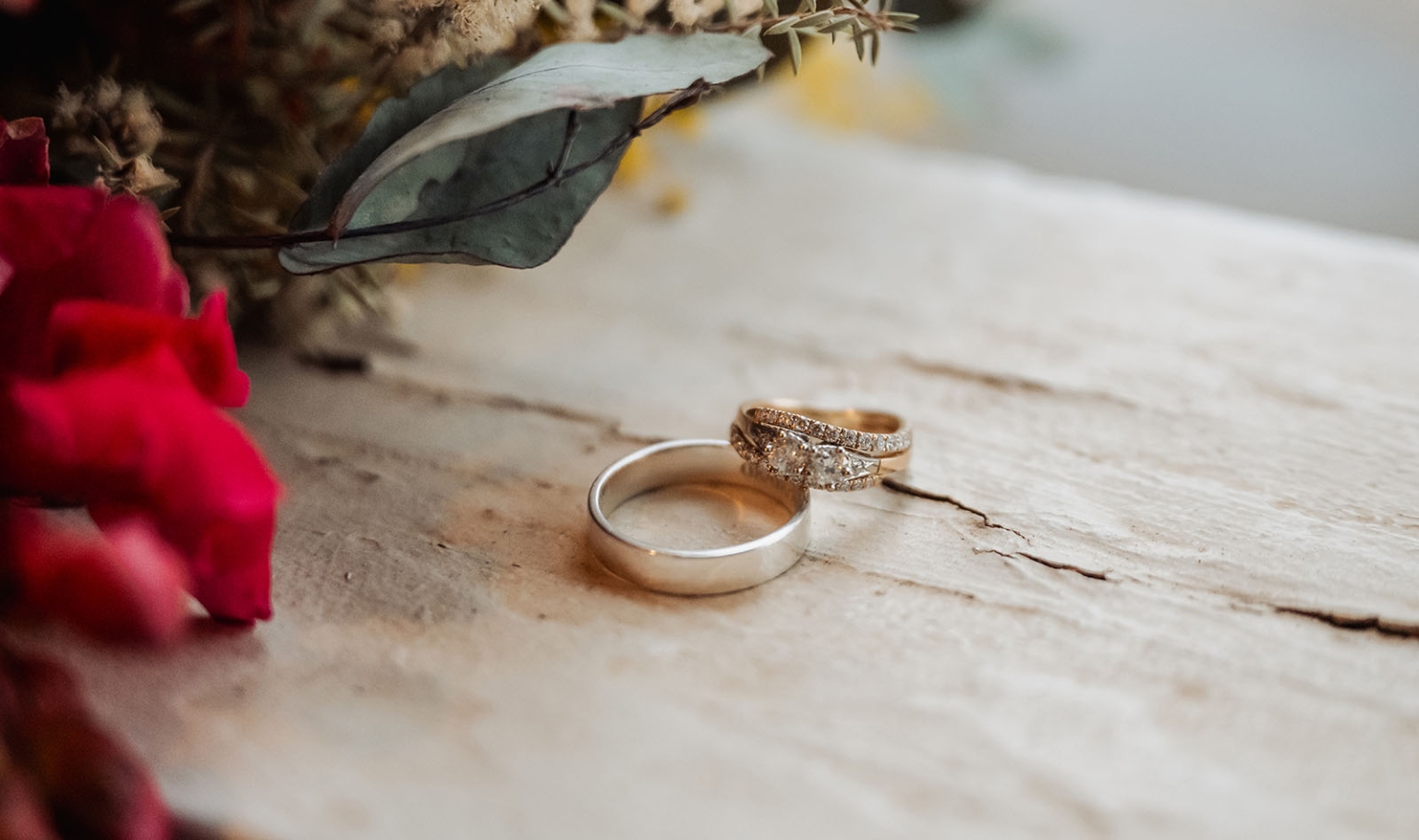 keyneton soldier's memorial hall wedding - lara & simon 44 Gold wedding bands resting beside red flowers and eucalyptus on a weathered timber surface