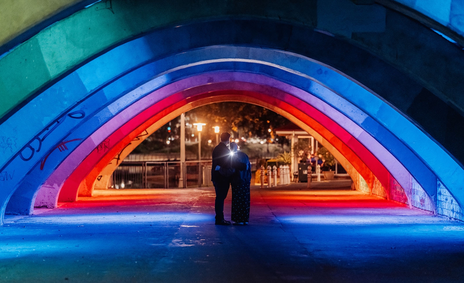 elder park rotunda proposal - lauren & enrique 11 Lauren and Enrique embracing beneath a brightly lit multicoloured concrete archway at night in Adelaide