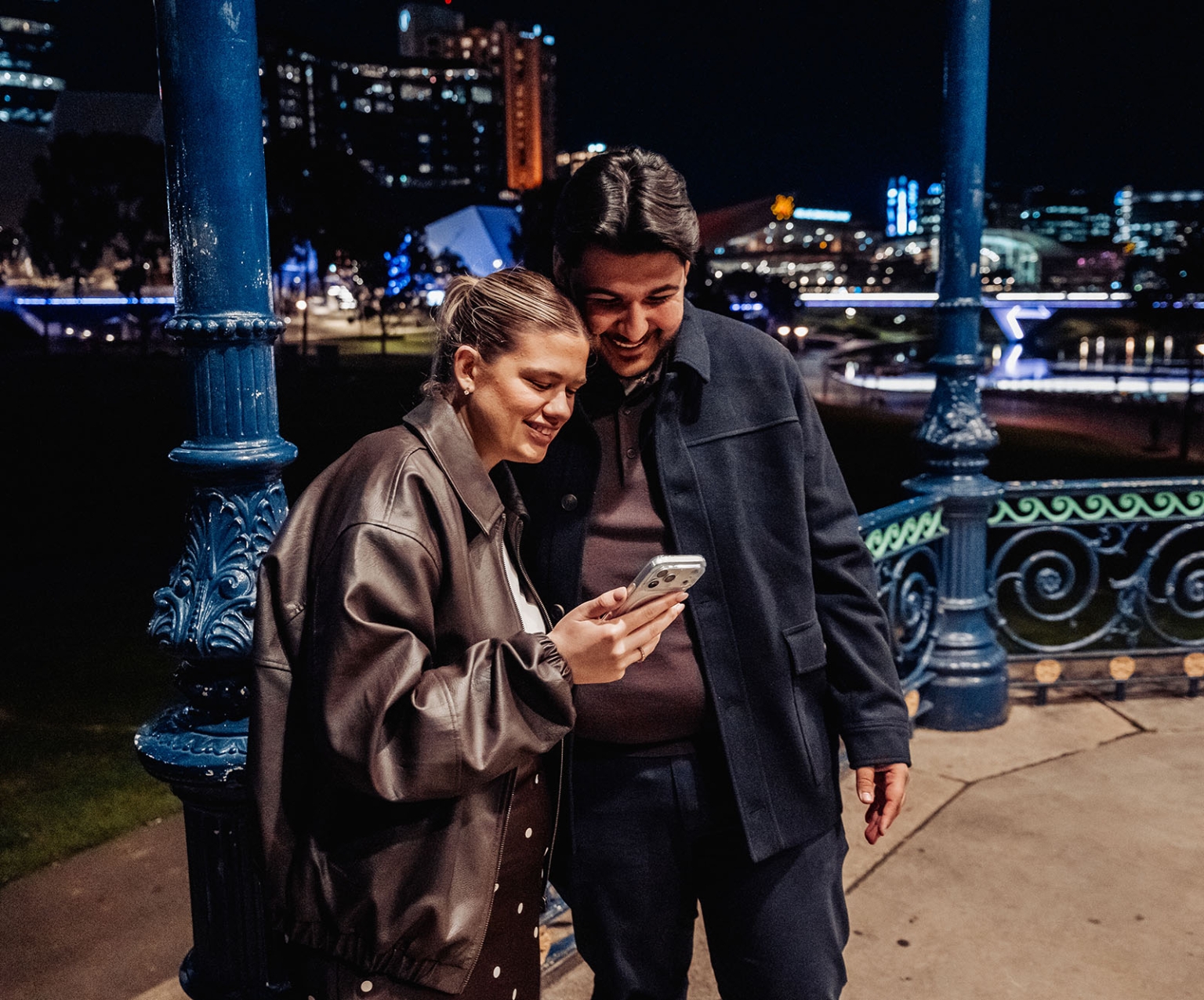 elder park rotunda proposal - lauren & enrique 6 Lauren and Enrique smiling together while looking at a phone beside the Elder Park Rotunda at night
