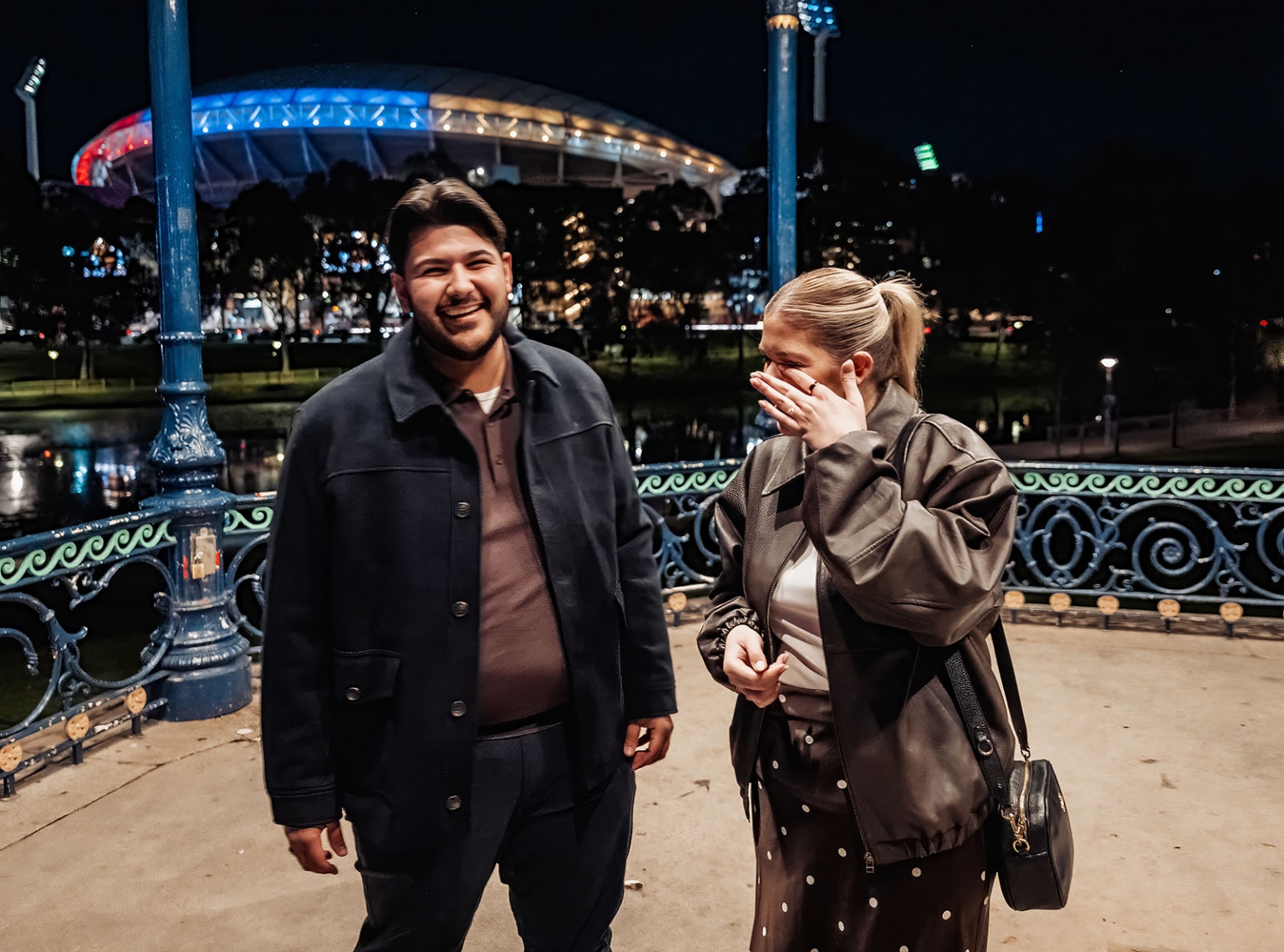 elder park rotunda proposal - lauren & enrique 4 Lauren covers her face as Enrique smiles beside her at Elder Park Rotunda at night, with Adelaide Oval lit behind them.