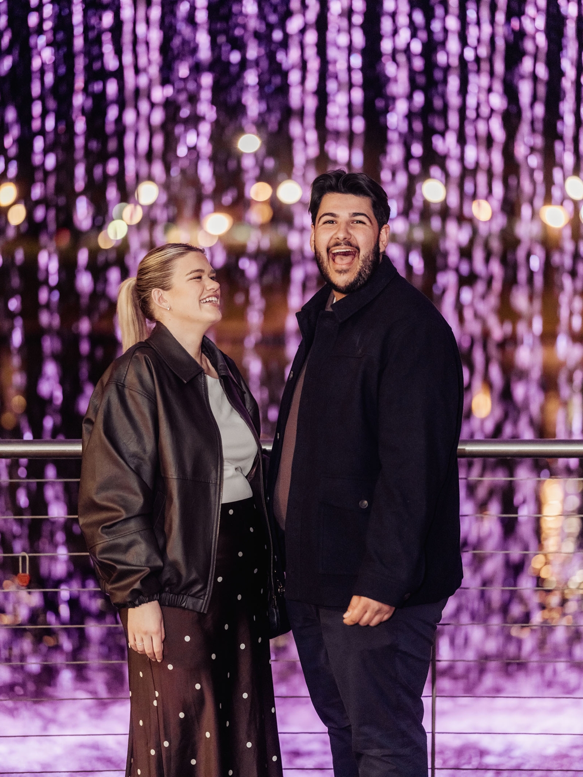 elder park rotunda proposal - lauren & enrique 13 Lauren and Enrique laughing together beside a railing with purple hanging lights behind them