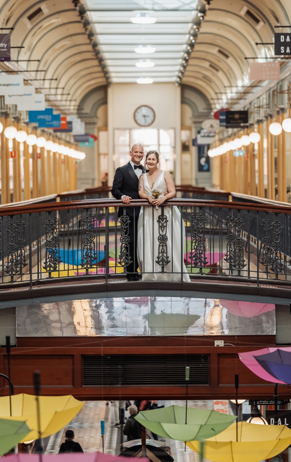 marriage registration office wedding - mia & roberto 101 Mia and Roberto in wedding attire standing on an ornate indoor arcade balcony, with colourful umbrellas below.