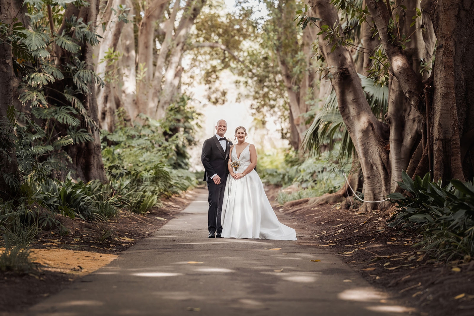 marriage registration office wedding - mia & roberto 59 Mia and Roberto standing together on a tree-lined path in Adelaide Botanic Park, with Mia in a white wedding gown holding a bouquet and Roberto in a black tuxedo