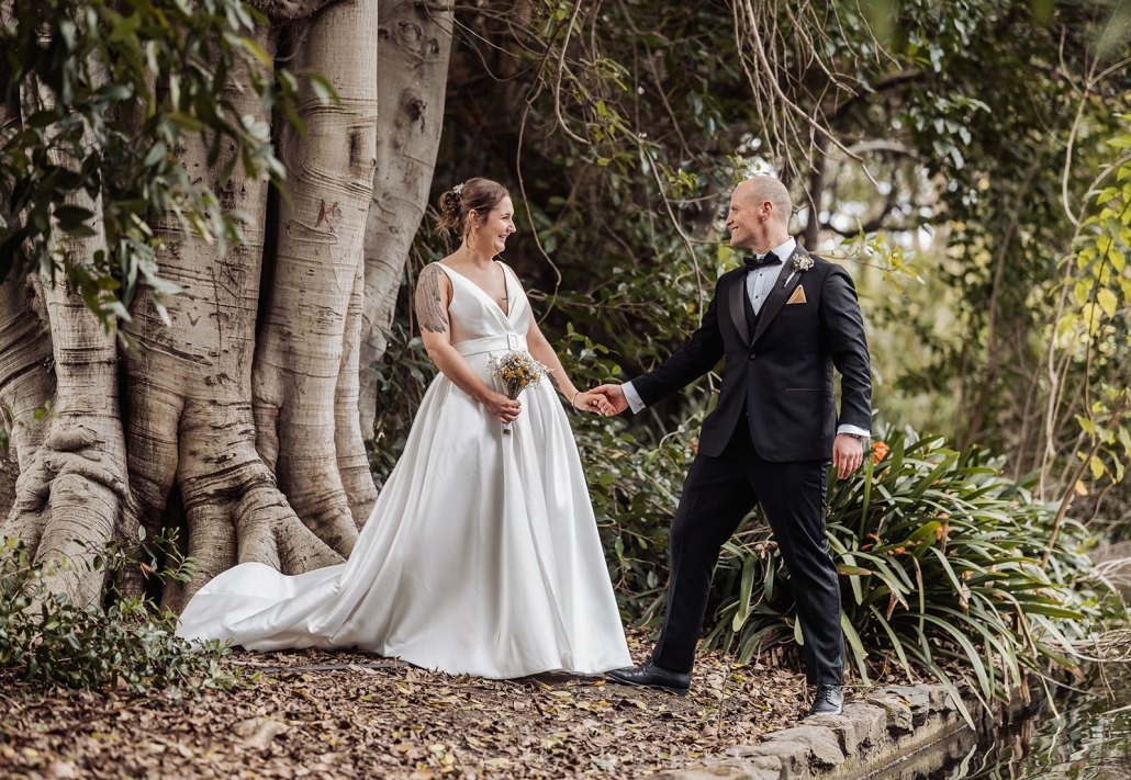 Mia and Roberto holding hands beside a large fig tree in Botanic Park, wearing a white wedding dress and black tuxedo.