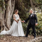 Mia and Roberto holding hands beside a large fig tree in Botanic Park, wearing a white wedding dress and black tuxedo.