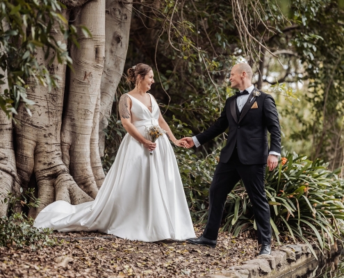 Mia and Roberto holding hands beside a large fig tree in Botanic Park, wearing a white wedding dress and black tuxedo.
