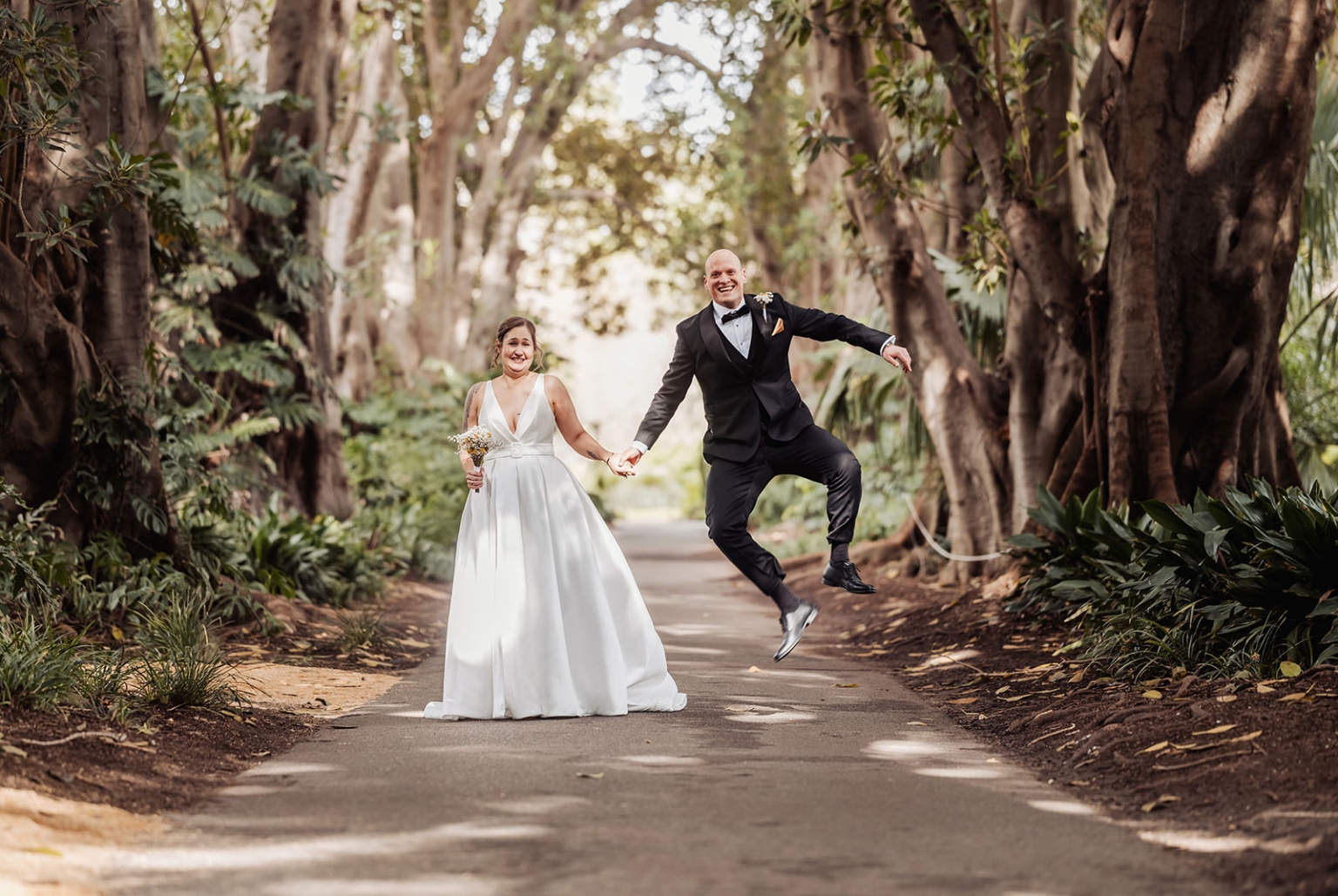 marriage registration office wedding - mia & roberto 61 Mia and Roberto holding hands on a tree-lined Botanic Park path, with Roberto jumping beside Mia in her white wedding dress.