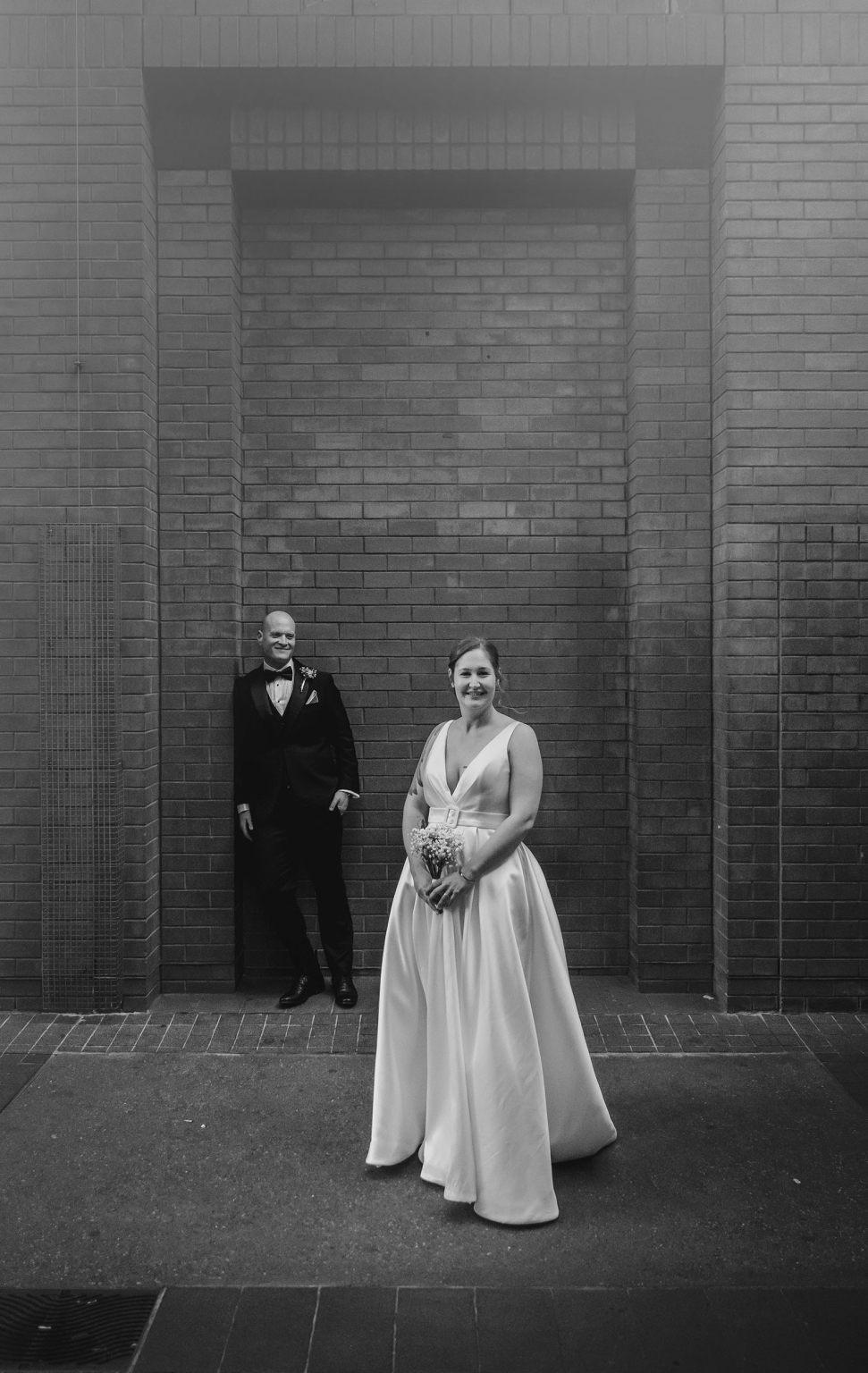 marriage registration office wedding - mia & roberto 94 Bride holding a small bouquet in a white wedding gown, with the groom in a black tuxedo behind her against a brick wall.