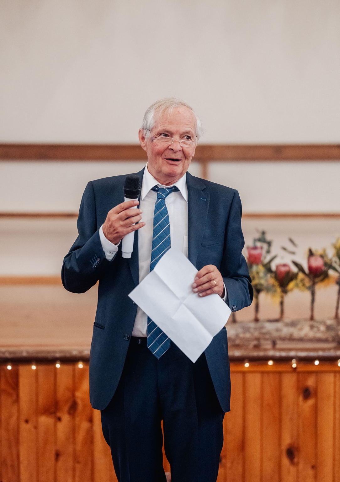 keyneton soldier's memorial hall wedding - lara & simon 43 Older man in a navy suit holding a microphone and notes while giving a wedding speech indoors