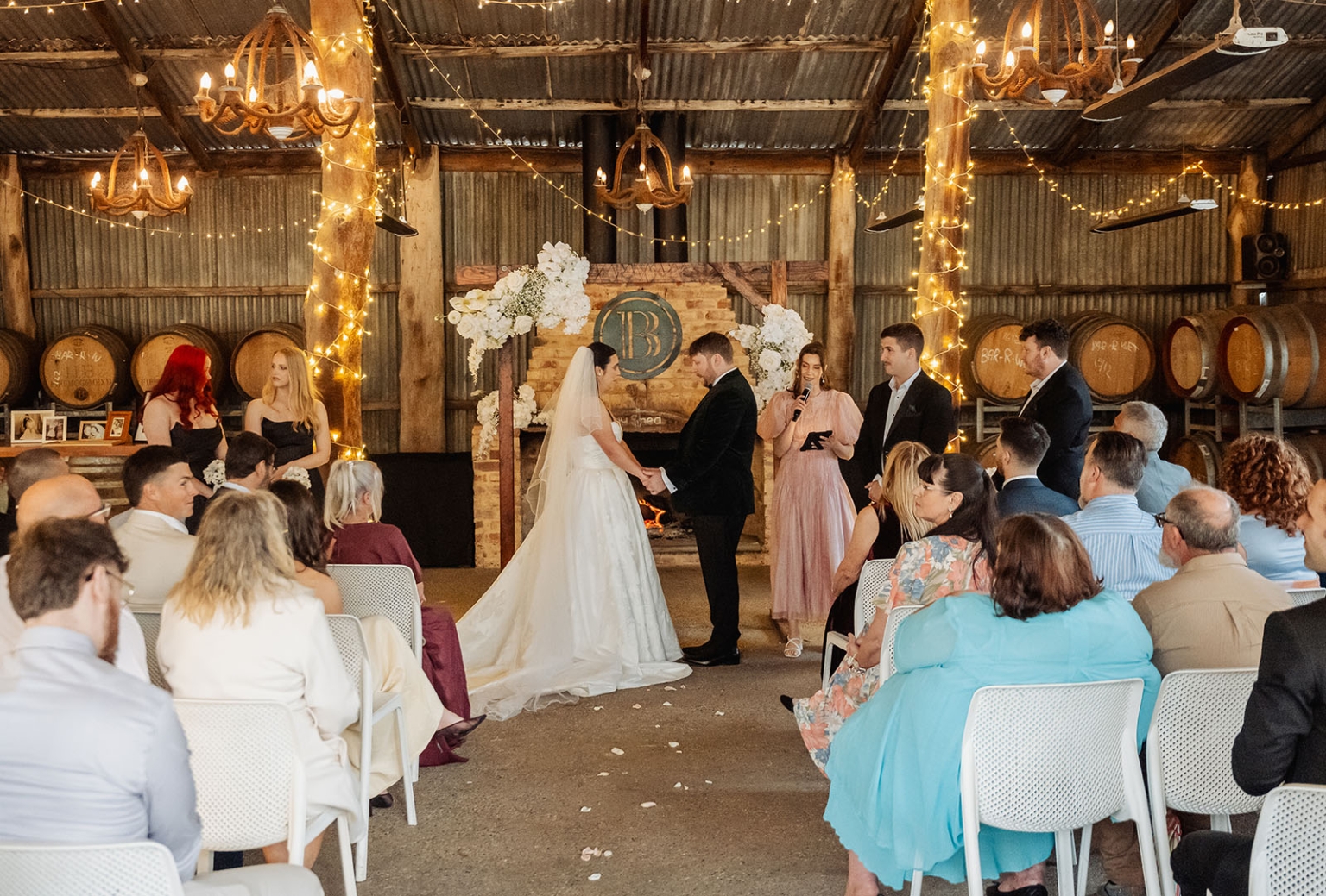 Bride and groom holding hands during a rustic indoor wedding ceremony at Barristers Block, surrounded by seated guests and fairy lights.