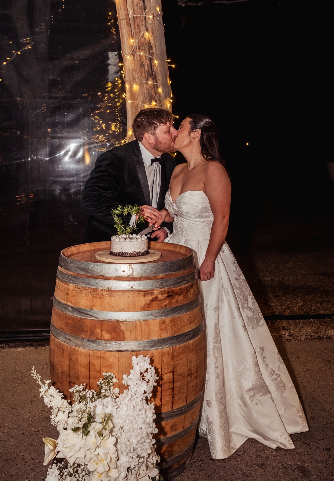 Newlyweds kissing beside their wedding cake on a wine barrel lit with fairy lights at Barristers Block.