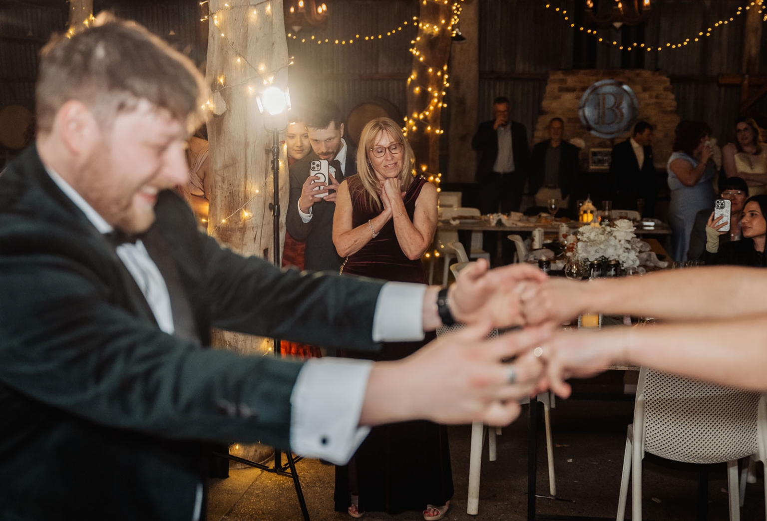 Groom in a tuxedo holding his partner’s hands while wedding guests watch beneath warm fairy lights at Barristers Block.
