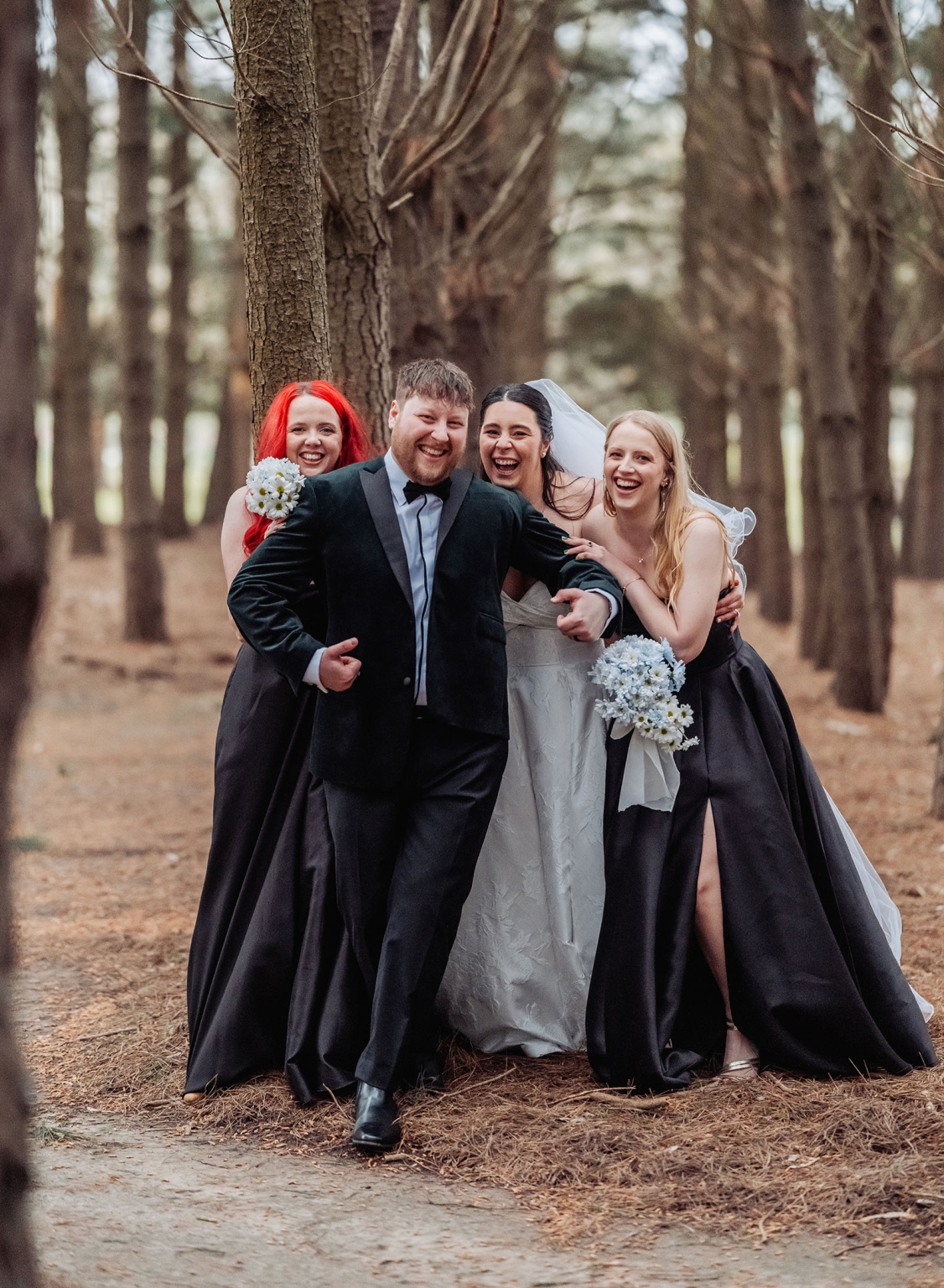 Bride, groom and two bridesmaids laughing together among tall trees, with the bridesmaids holding white flower bouquets.