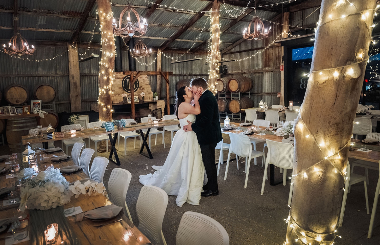 Bride and groom kissing between set reception tables in a rustic, fairy-lit barn at Barristers Block