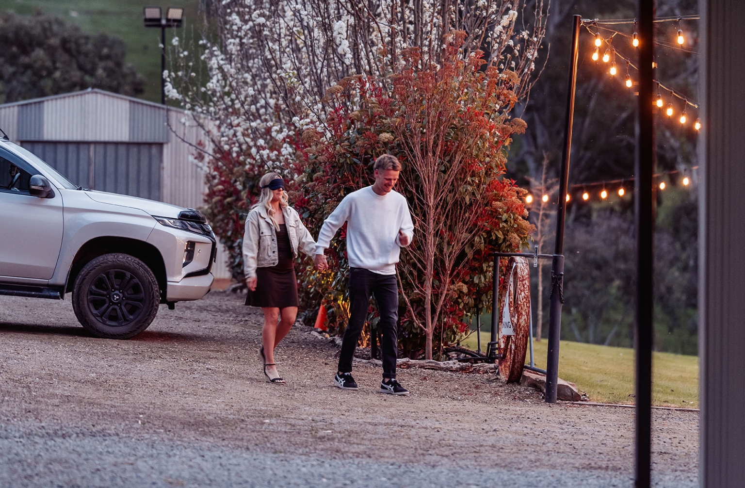 tiny house on the prairie proposal - kyle & bianca 2 A man leads a blindfolded woman by the hand along a gravel path beneath fairy lights at Tiny House on the Prairie.
