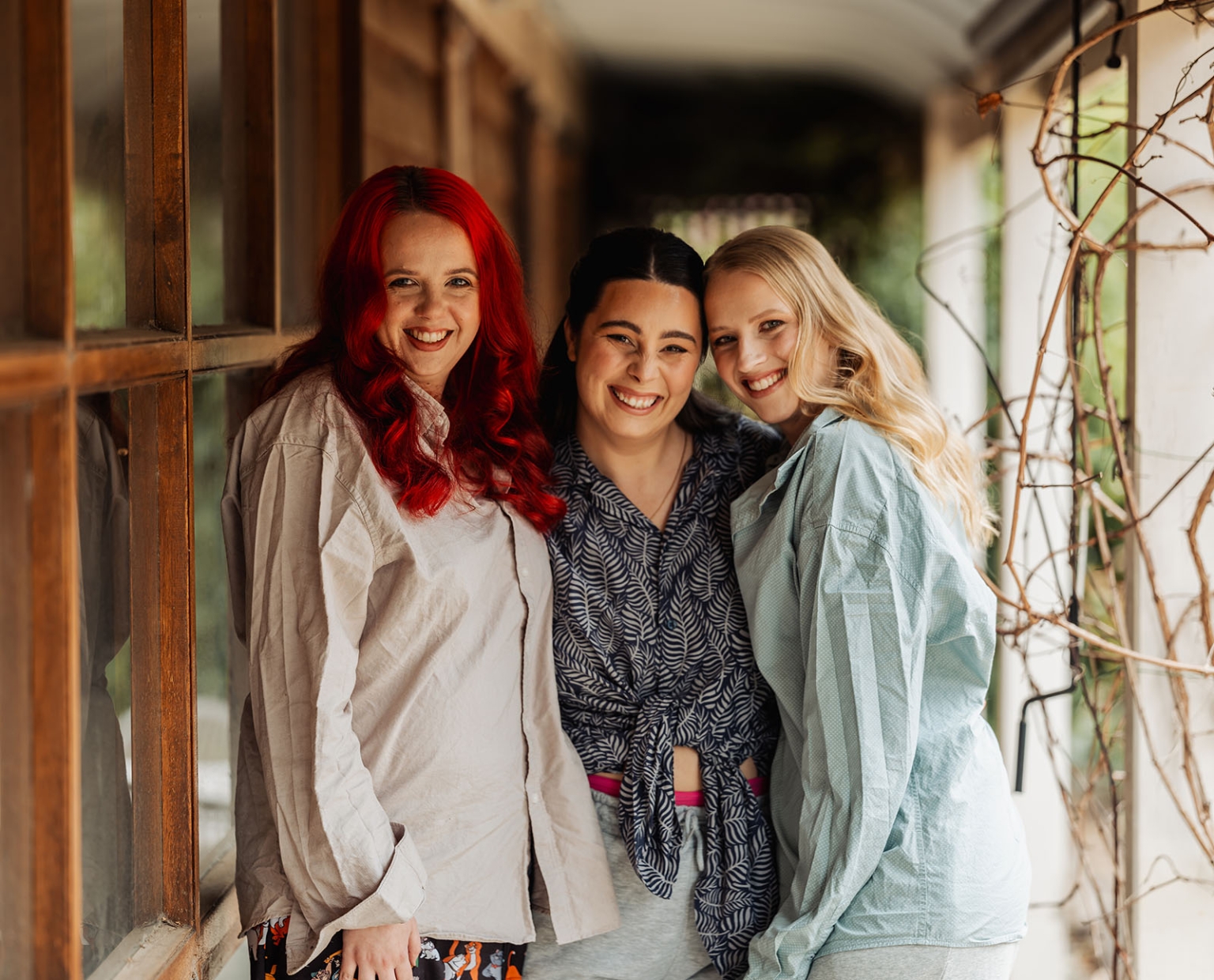 Three women in casual clothes smiling together beside a timber-framed window during wedding preparations near Mount Barker.
