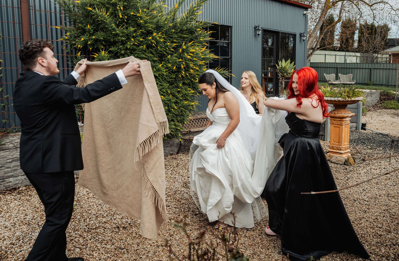 Bride in a white wedding gown walking outdoors as a man holds a blanket and bridesmaids help with her dress