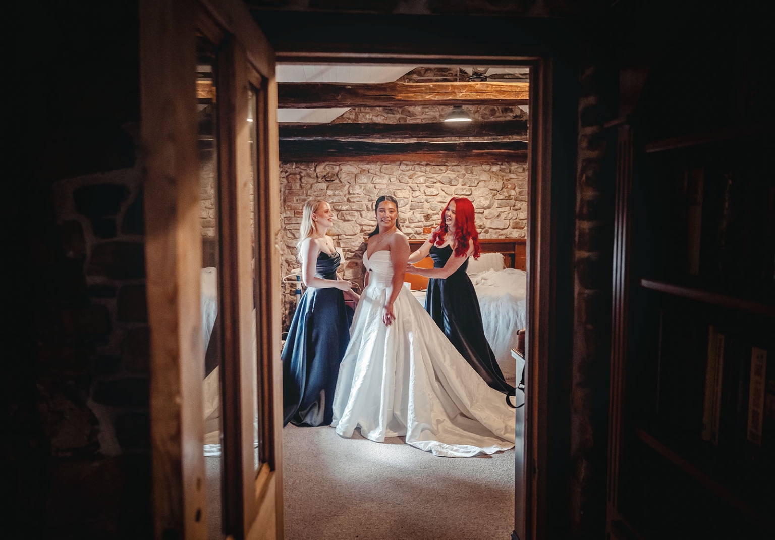 Bride in a white wedding gown standing with two bridesmaids in dark dresses inside a rustic stone-walled cottage bedroom