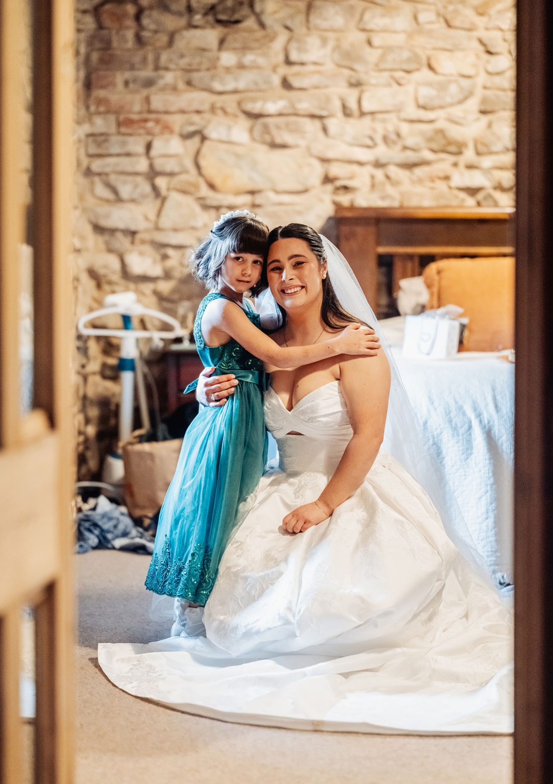 Bride in a white wedding gown kneeling beside a young girl in a teal dress inside a rustic stone-walled cottage.