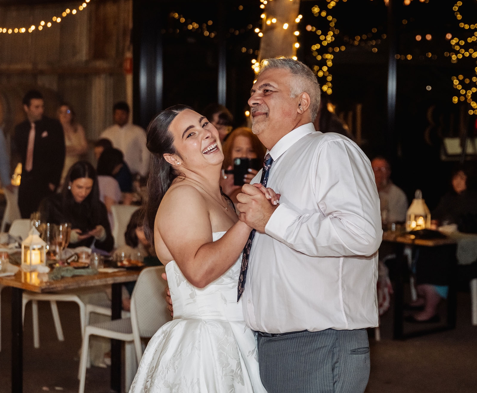 Bride in a white wedding dress dancing with a man at a warmly lit reception, surrounded by seated guests and fairy lights.