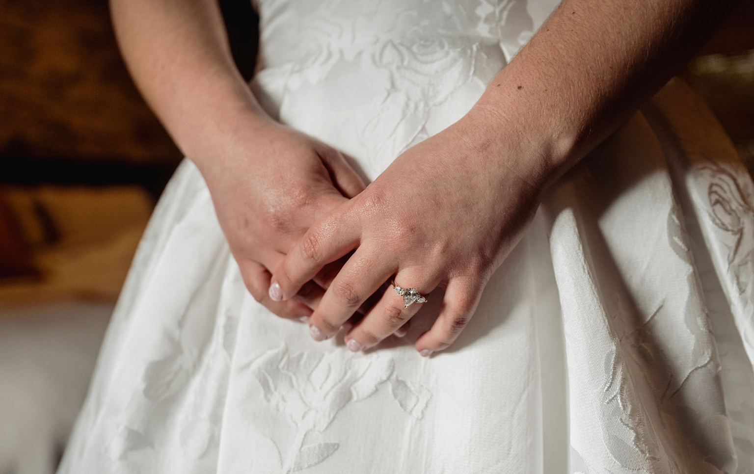 Bride's hands resting on her embroidered white wedding dress, showing a diamond engagement ring.