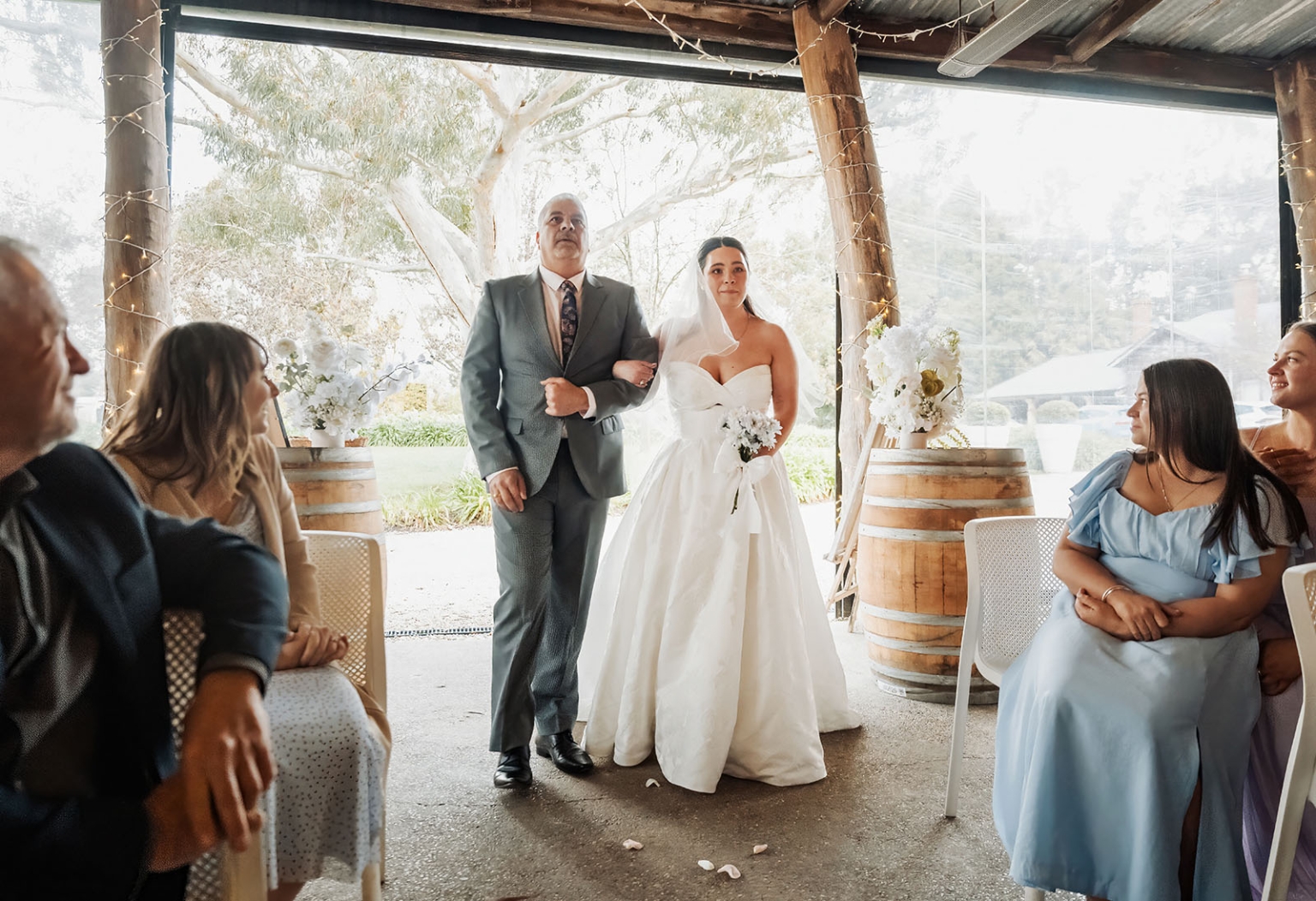Bride in a strapless white wedding gown walks arm in arm with her father into a rustic ceremony space as guests watch.