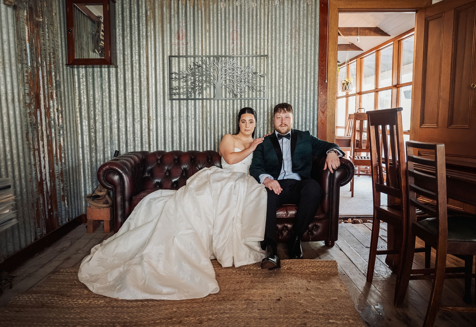 Bride and groom seated together on a brown leather sofa in a rustic cottage with corrugated iron walls and timber furniture