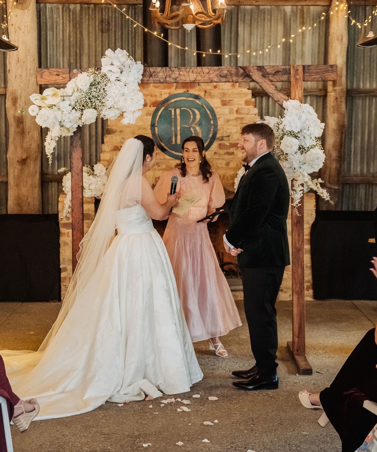 Carley and Corey smiling with their celebrant beneath a white floral wedding arbour at Barristers Block
