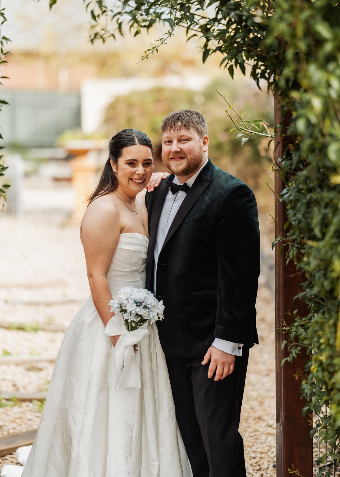 Carley and Corey smiling together in wedding attire beneath leafy garden foliage at Barristers Block