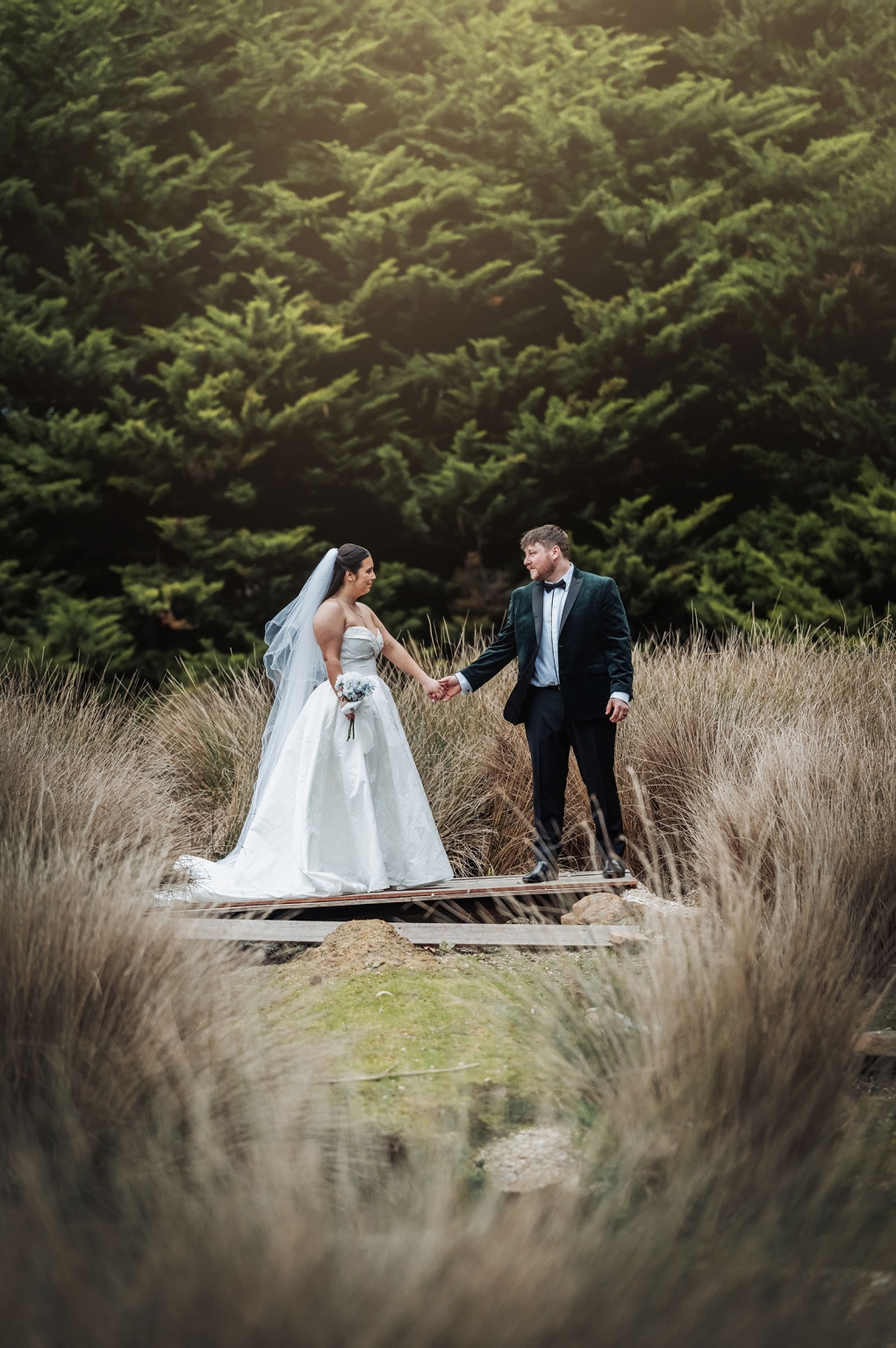 Carley and Corey holding hands on a small timber bridge, surrounded by tall grass and dense ferns at Barristers Block