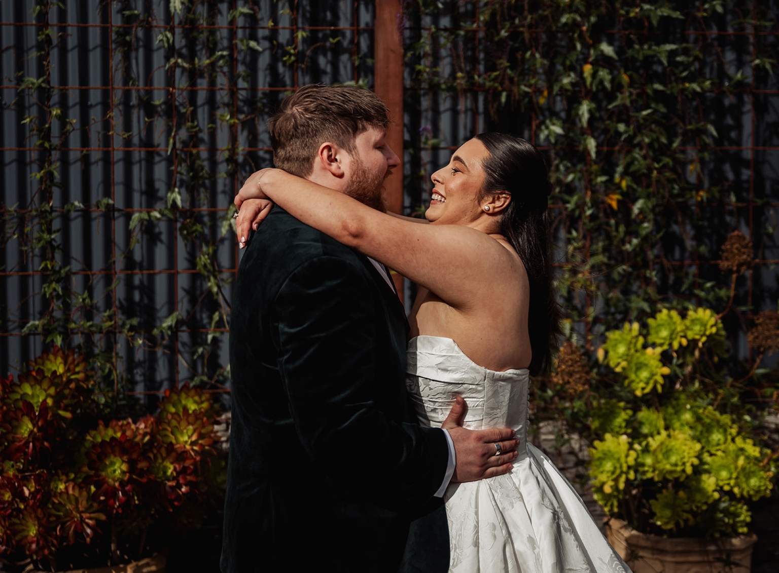 Carley and Corey smiling as they embrace in a leafy garden, with Carley wearing a white wedding dress and Corey a dark suit jacket.