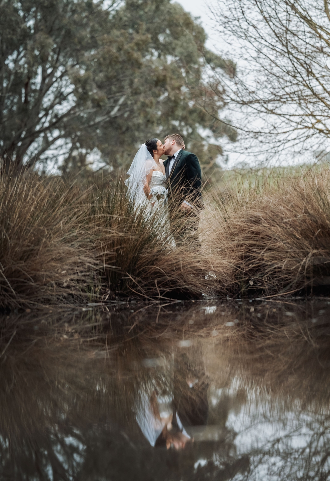 Carley and Corey kissing beside a pond, with their reflection in the water and tall grass surrounding them